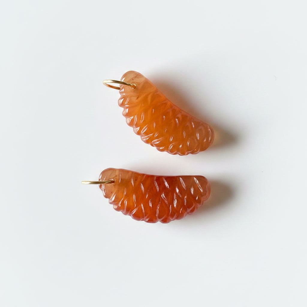 Four hand-carved gemstone charms shaped like citrus slices—two in carnelian and two in lemon quartz—with 14k gold rings, displayed on dark gray fabric.